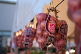 Bags of red onions are hanging, organized in a row, likely at a market stall. The onions are enclosed in mesh bags with visible labels that display information and branding. A soft focus in the background suggests other similar bags displayed alongside.