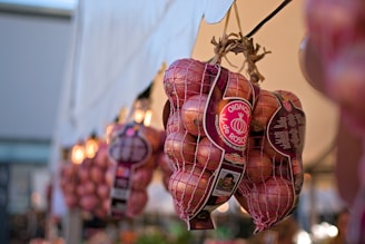 Bags of red onions are hanging, organized in a row, likely at a market stall. The onions are enclosed in mesh bags with visible labels that display information and branding. A soft focus in the background suggests other similar bags displayed alongside.
