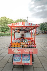 A street vendor cart loaded with various colorful bottled drinks is positioned on a paved area. The vendor is partially visible behind the cart, under a canopy with bright signage. The bottles are neatly arranged in rows displaying different brands and flavors. Trees and other urban elements are visible in the background under a cloudy sky.