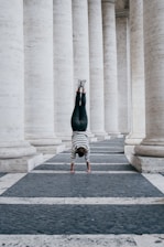 A person performs a handstand in a corridor lined with large stone columns. The individual is wearing a striped shirt and dark pants, with feet elevated against the backdrop of the architectural structure.