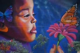 A vibrant mural of a child with butterflies, featuring a close-up of a child facing a large butterfly perched on a pink flower. The child appears calm and contemplative, with one blue butterfly resting on their head. The background is a deep blue, enhancing the colors of the butterflies and flower.