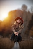 A woman wearing a warm hat and sweater is sitting on a railroad track with grass around her. She appears relaxed and is smiling softly. The background is blurred, with an orange sunset glow providing a warm atmosphere.
