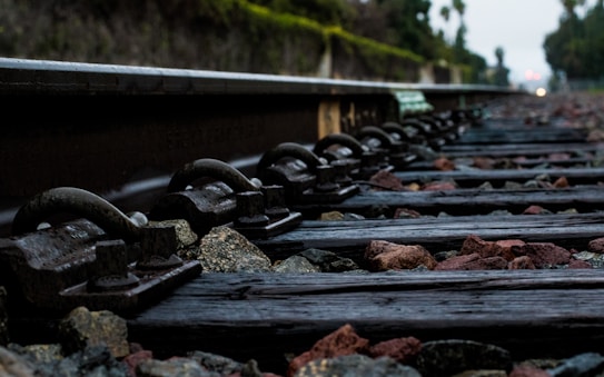 A close-up view of railway tracks with wooden sleepers and scattered stones between them. The image is taken from a low angle, emphasizing the texture of the metal rails and the earthy tones of the stones. In the background, there are blurred trees and greenery, suggesting an outdoor setting.