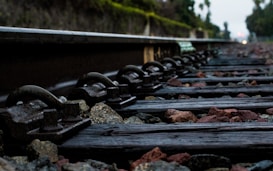 A close-up view of railway tracks with wooden sleepers and scattered stones between them. The image is taken from a low angle, emphasizing the texture of the metal rails and the earthy tones of the stones. In the background, there are blurred trees and greenery, suggesting an outdoor setting.