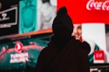 A silhouette of a person wearing a beanie is seen against a backdrop of brightly lit, colorful billboards with various advertisements, including recognizable brand logos.
