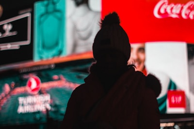 A silhouette of a person wearing a beanie is seen against a backdrop of brightly lit, colorful billboards with various advertisements, including recognizable brand logos.