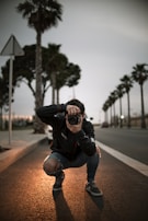 A person is crouching on a dimly lit street, holding a camera to their face while taking a photograph. The scene is framed with tall palm trees lining the road, and there is a moody, overcast sky. The lighting creates a warm glow on the asphalt around the person, emphasizing a focus on the photographer.