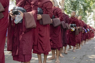 A group of monks wearing deep maroon robes stand in a single file line on a tree-lined street. Each monk holds a black alms bowl, and some also carry items such as plastic cups and bottles. The monks appear to be barefoot, and they are positioned closely together in a calm and orderly formation.