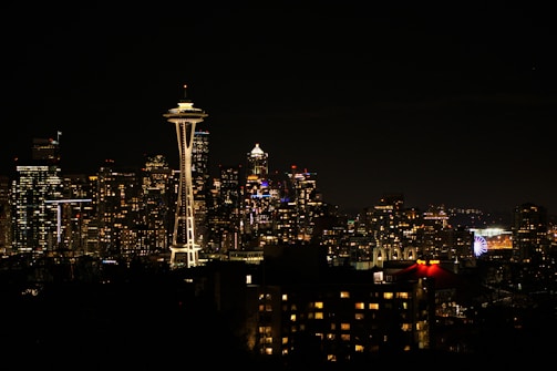 A nighttime view of a city skyline with numerous illuminated skyscrapers and buildings. The prominent Space Needle stands out, glowing against the dark sky. A Ferris wheel is visible in the distance, adding to the urban landscape.
