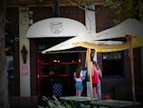 A street view of a brick building with a 'Burrito' sign above the entrance. Two women are standing outside under a large Corona-branded patio umbrella, looking at a menu or sign. String lights are seen above the entrance, and there is greenery in the foreground.