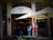 A street view of a brick building with a 'Burrito' sign above the entrance. Two women are standing outside under a large Corona-branded patio umbrella, looking at a menu or sign. String lights are seen above the entrance, and there is greenery in the foreground.