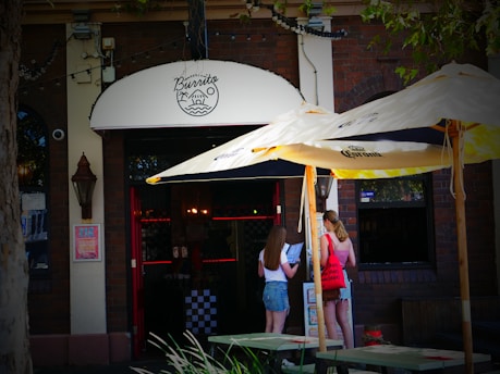A street view of a brick building with a 'Burrito' sign above the entrance. Two women are standing outside under a large Corona-branded patio umbrella, looking at a menu or sign. String lights are seen above the entrance, and there is greenery in the foreground.