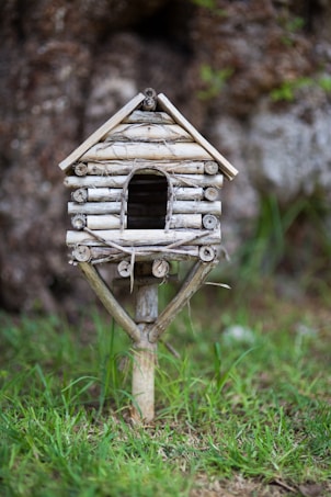 A small, handcrafted birdhouse made of twigs and natural materials stands on a wooden post amidst a grassy area. The birdhouse has a rustic appearance with a simple design, including a pitched roof and an arched entrance. The background features a blurred natural setting with hints of greenery and tree bark.