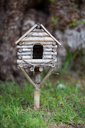 A small, handcrafted birdhouse made of twigs and natural materials stands on a wooden post amidst a grassy area. The birdhouse has a rustic appearance with a simple design, including a pitched roof and an arched entrance. The background features a blurred natural setting with hints of greenery and tree bark.