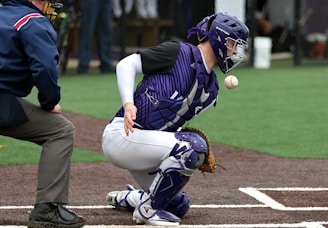 A baseball catcher in purple and white gear is crouched down, focused on an incoming ball. The umpire, positioned close to the catcher, is observing the play. The field is artificial turf with a dirt-colored infield, and blurred figures are in the background.