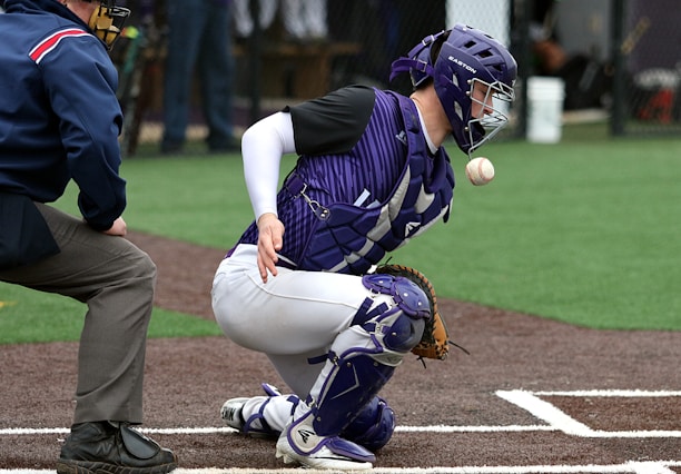A baseball catcher in purple and white gear is crouched down, focused on an incoming ball. The umpire, positioned close to the catcher, is observing the play. The field is artificial turf with a dirt-colored infield, and blurred figures are in the background.