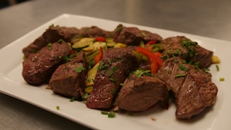 A plate featuring several cuts of grilled steak garnished with fresh herbs, accompanied by sliced grilled vegetables including zucchini and red peppers, arranged on a white rectangular plate.