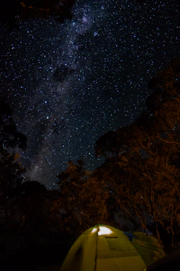 A star-filled night sky above a quiet campsite, with tents softly lit from within.