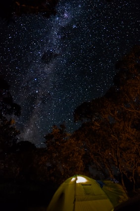 A star-filled night sky with the Milky Way visible across the center. In the foreground, a warmly lit tent sits surrounded by silhouetted trees, creating a peaceful and serene outdoor setting.