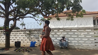 A woman in a patterned red dress walks past a display of sandals under a large tree. Various sandals are laid out on the ground in front of the tree, and bags are placed nearby. In the background, a man wearing a hat sits against a white wall topped with barbed wire. The scene is set outdoors, with a partly cloudy sky.