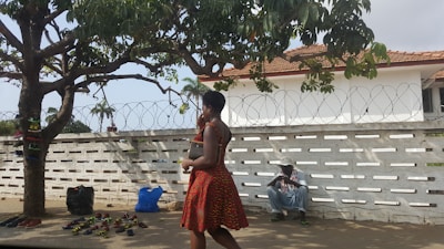 A woman in a patterned red dress walks past a display of sandals under a large tree. Various sandals are laid out on the ground in front of the tree, and bags are placed nearby. In the background, a man wearing a hat sits against a white wall topped with barbed wire. The scene is set outdoors, with a partly cloudy sky.