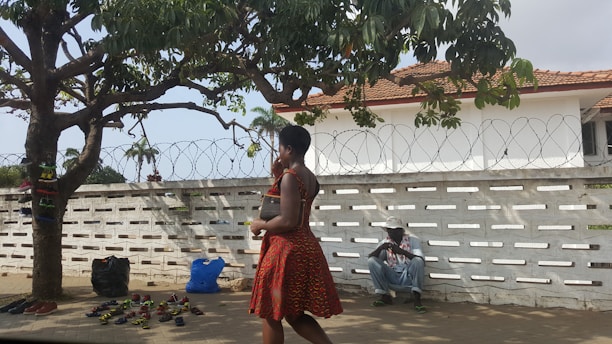 A woman in a patterned red dress walks past a display of sandals under a large tree. Various sandals are laid out on the ground in front of the tree, and bags are placed nearby. In the background, a man wearing a hat sits against a white wall topped with barbed wire. The scene is set outdoors, with a partly cloudy sky.
