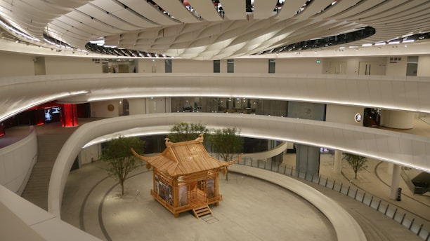 A futuristic interior space featuring a central wooden gazebo-like structure surrounded by curved architectural elements and an atrium. The ceiling design is intricate and modern, with a mix of white and metallic tones. There are a few small trees placed around the structure, adding a touch of nature to the otherwise sleek and minimalistic design.