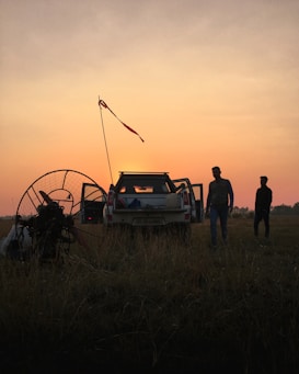 A scenic landscape at sunset with silhouettes of two people near an open vehicle in a field. The sky is a gradient of orange and pink, creating a calming backdrop. A paramotor is visible on the left side, indicating preparations or activities related to paragliding.