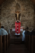 A priest in a red robe stands before an altar in a stone-walled church, holding a chalice. A crucifix with Jesus is mounted on the wall above the altar. Several people are seated in the pews, observing the ceremony.