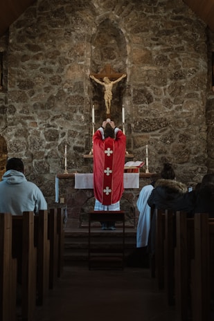 A priest in a red robe stands before an altar in a stone-walled church, holding a chalice. A crucifix with Jesus is mounted on the wall above the altar. Several people are seated in the pews, observing the ceremony.