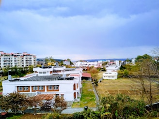 A suburban area featuring a variety of buildings under a blue sky. The foreground includes a white building with a red roof surrounded by greenery and neatly trimmed bushes. In the background, apartment complexes and smaller residential buildings are visible, with a mix of trees and open fields around them. The overall setting is a mix of urban and natural elements.