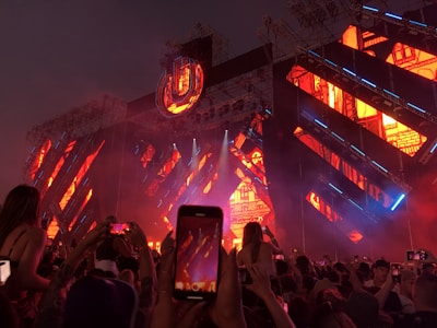 A vibrant live concert or festival setting with a large crowd of people raising their phones to capture the moment. The stage is illuminated with bright orange, blue, and purple lights with intricate patterns. An ornate, illuminated sign or logo is prominently displayed above the stage.
