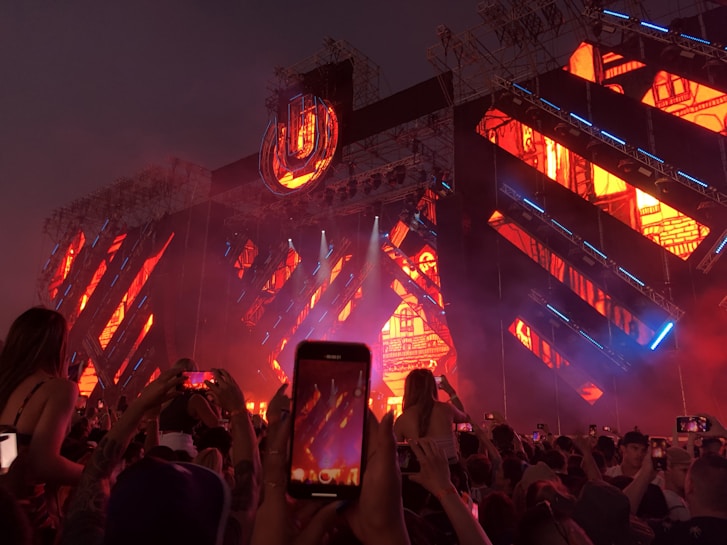 A vibrant live concert or festival setting with a large crowd of people raising their phones to capture the moment. The stage is illuminated with bright orange, blue, and purple lights with intricate patterns. An ornate, illuminated sign or logo is prominently displayed above the stage.