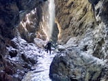 A person wearing a helmet and protective gear navigates through a narrow, rocky canyon with high walls. Sunlight filters through, creating bright patches and highlighting the rugged textures of the rock. Water flows along the uneven floor, adding a reflective sheen to the scene.