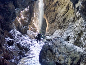A person wearing a helmet and protective gear navigates through a narrow, rocky canyon with high walls. Sunlight filters through, creating bright patches and highlighting the rugged textures of the rock. Water flows along the uneven floor, adding a reflective sheen to the scene.