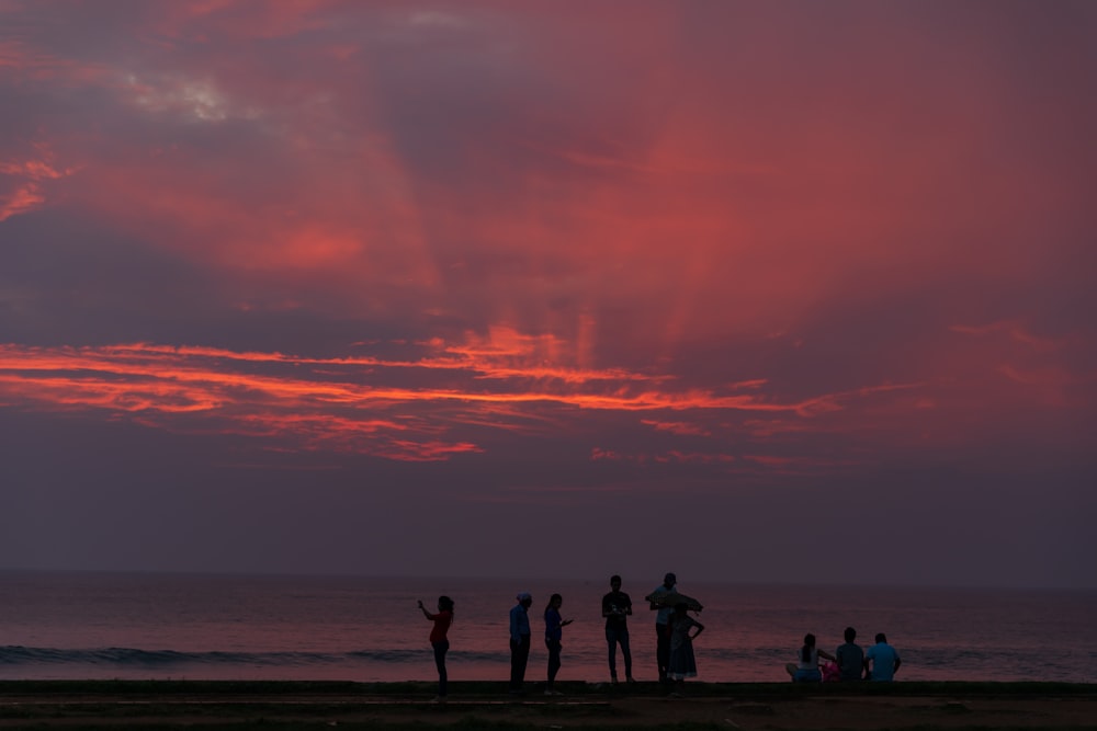Silueta de personas de pie y sentadas cerca de la playa durante la puesta del sol