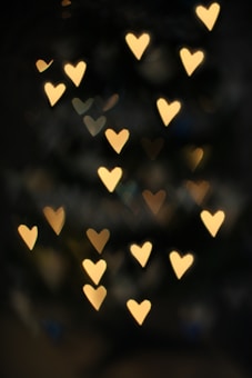 A dark background is punctuated by numerous glowing heart-shaped lights in various shades of yellow and gold, creating a soft and romantic atmosphere.