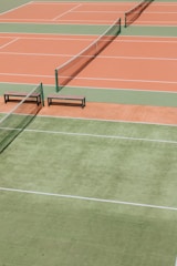 Several tennis courts with orange and green surfaces, separated by nets. Benches are placed alongside the courts.