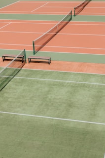 Several tennis courts with orange and green surfaces, separated by nets. Benches are placed alongside the courts.