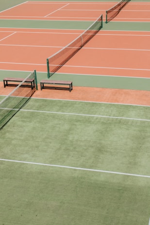 Several tennis courts with orange and green surfaces, separated by nets. Benches are placed alongside the courts.