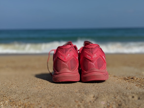 Red athletic shoes placed on a sandy beach with ocean waves in the background. The scene implies a leisurely atmosphere and a connection to nature and relaxation.