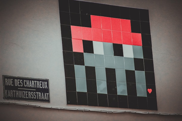 A pixel art piece on a wall depicting what appears to be a simple, stylized character or object using a blocky grid design. The artwork prominently features red, black, and gray tiles. A small heart shape is visible in the lower right corner of the art. Beside it, a street sign in French and Dutch reads 'RUE DES CHARTREUX' and 'KARTHUIZERSSTRAAT'.