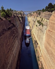 A large cargo ship navigates through a narrow canal, flanked on both sides by steep rock walls. A metal bridge spans across the top of the canal, surrounded by lush green trees.