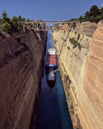 A large cargo ship navigates through a narrow canal, flanked on both sides by steep rock walls. A metal bridge spans across the top of the canal, surrounded by lush green trees.