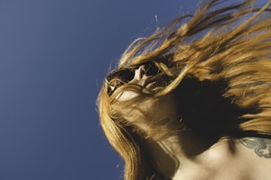 A person with long, flowing hair wears sunglasses against a clear blue sky. The wind sweeps the hair dramatically across the face, partially obscuring it. A tattoo is visible on the shoulder of the sunlit skin.