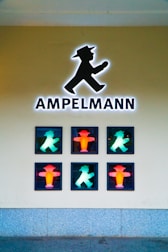 A display features the iconic pedestrian traffic light symbol known as Ampelmann, with six illuminated figures in a grid formation. The silhouettes are a mix of red and green, representing stop and go signals. The wall behind is plain and the floor appears to be paved.