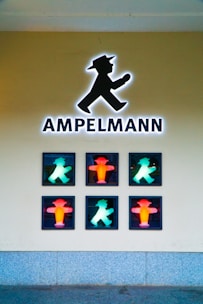 A display features the iconic pedestrian traffic light symbol known as Ampelmann, with six illuminated figures in a grid formation. The silhouettes are a mix of red and green, representing stop and go signals. The wall behind is plain and the floor appears to be paved.