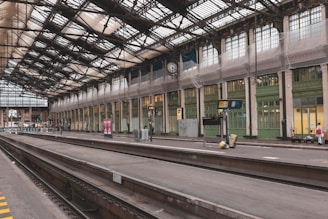 A spacious and historic railway station platform with intricate metal and glass roofing. The platform is mostly empty, with some luggage and a few people in the distance. There are large vintage clocks and electronic display boards. The station's architecture is classic, with high ceilings and ornamental details.