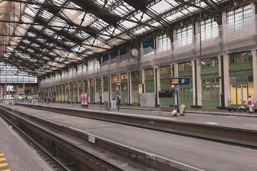 A spacious and historic railway station platform with intricate metal and glass roofing. The platform is mostly empty, with some luggage and a few people in the distance. There are large vintage clocks and electronic display boards. The station's architecture is classic, with high ceilings and ornamental details.