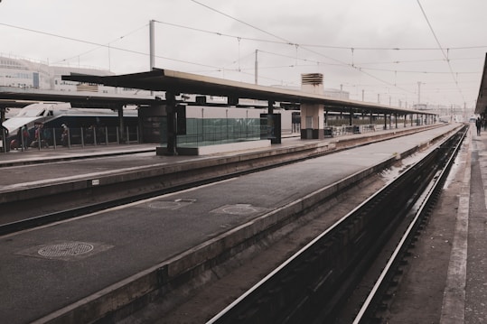 A train platform with an empty railway track. The platform is covered and features some glass barriers along the side. In the background, there is a high-speed train visible with a few people standing near it. Overhead, there are electric wires, and the sky is overcast.